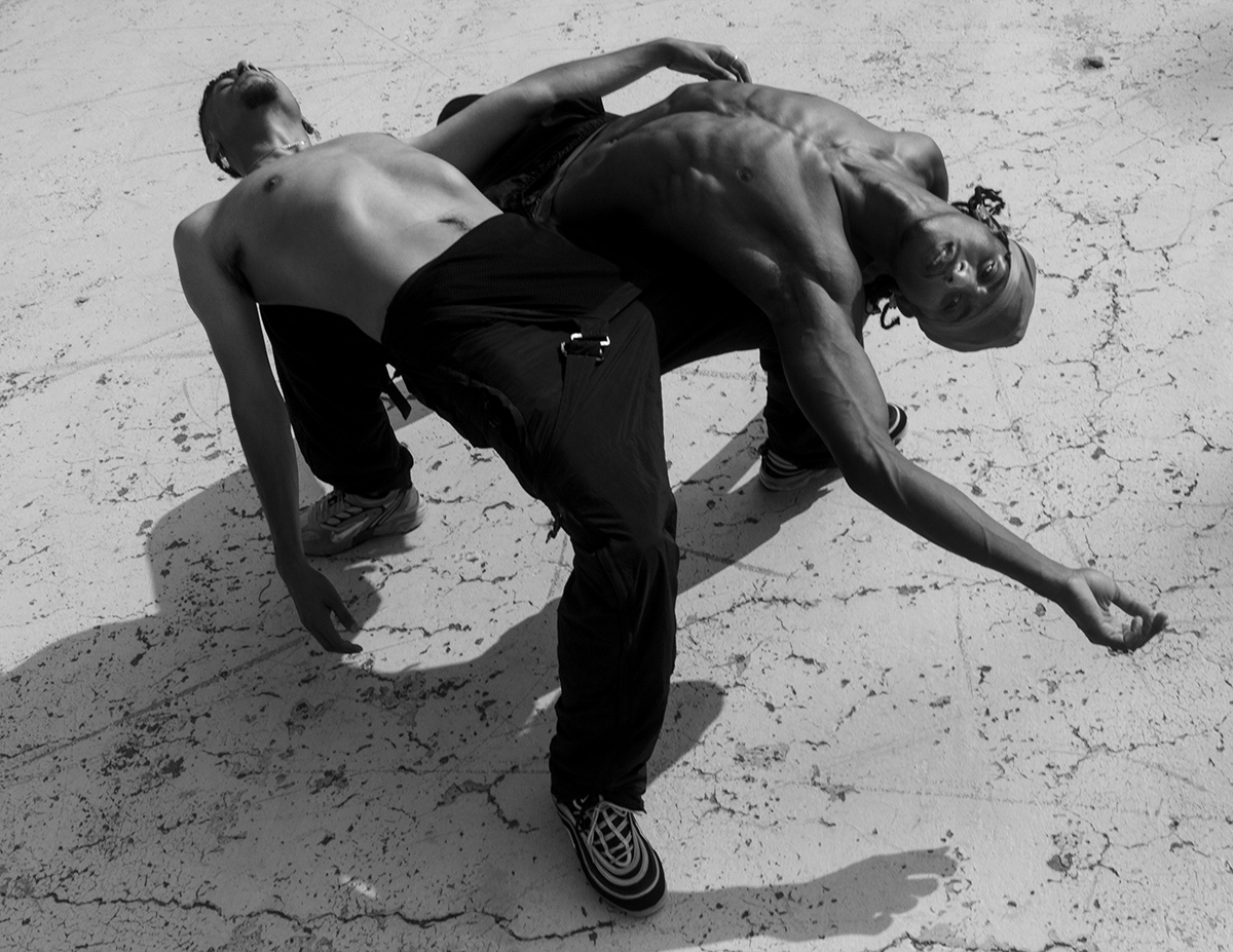 Photo of two Black men, both shirtless, bending backwards and leaning on each other's knees.