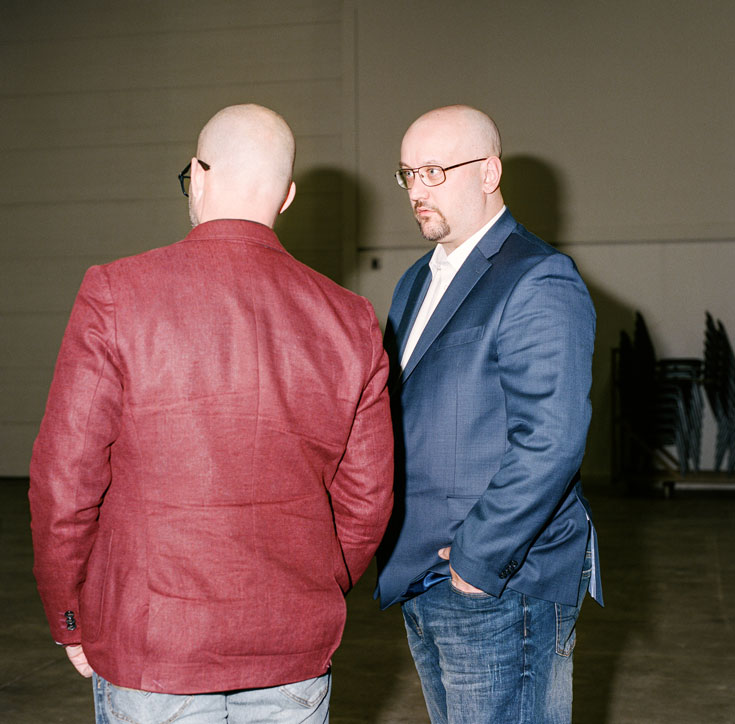 Two bald men are in conversation, one with his back to the camera wearing a red jacket. The other has a goatee and is wearing a blue blazer and jeans.