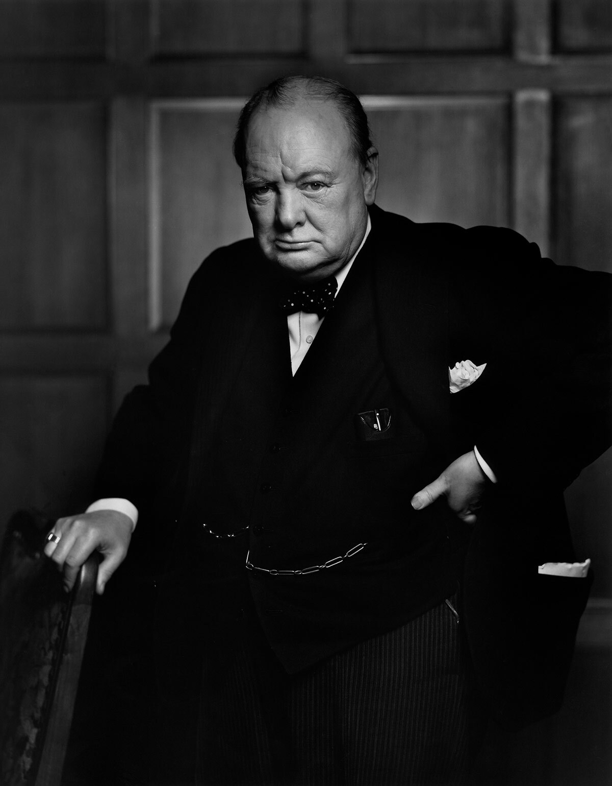 A black-and-white photo of Winston Churchill scowling and leaning his hand against a chair back