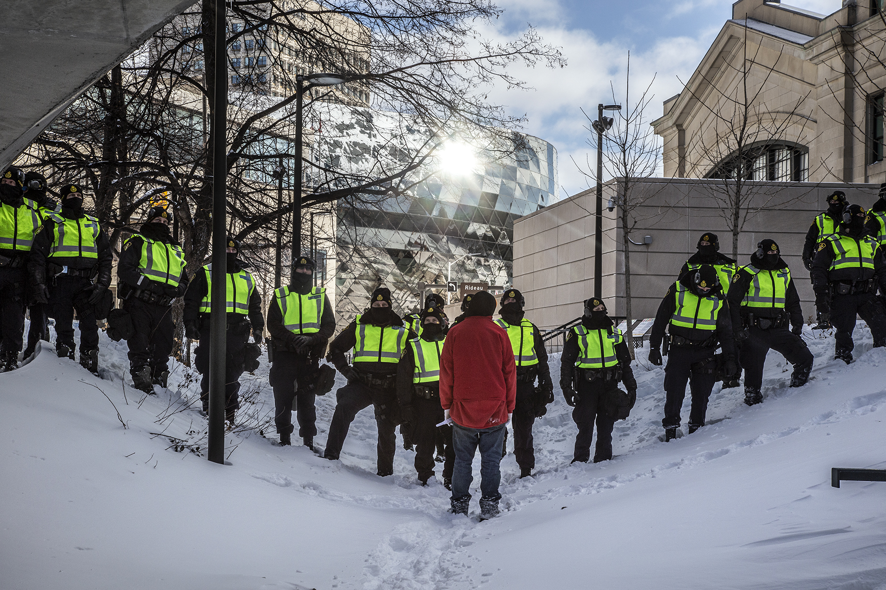A protester in a read coat stands before a line of police in neon vests in Ottawa