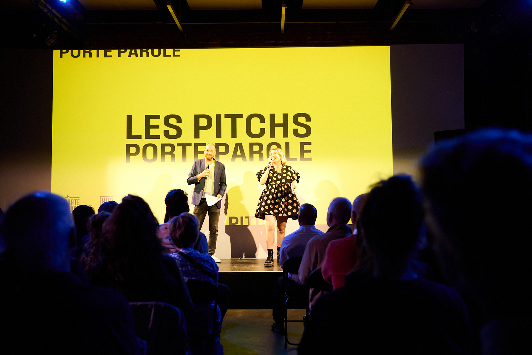 Photo of a man and a woman on a small stage in front of an audience. The background behind them is a plain, bright yellow and features bold black text reading 