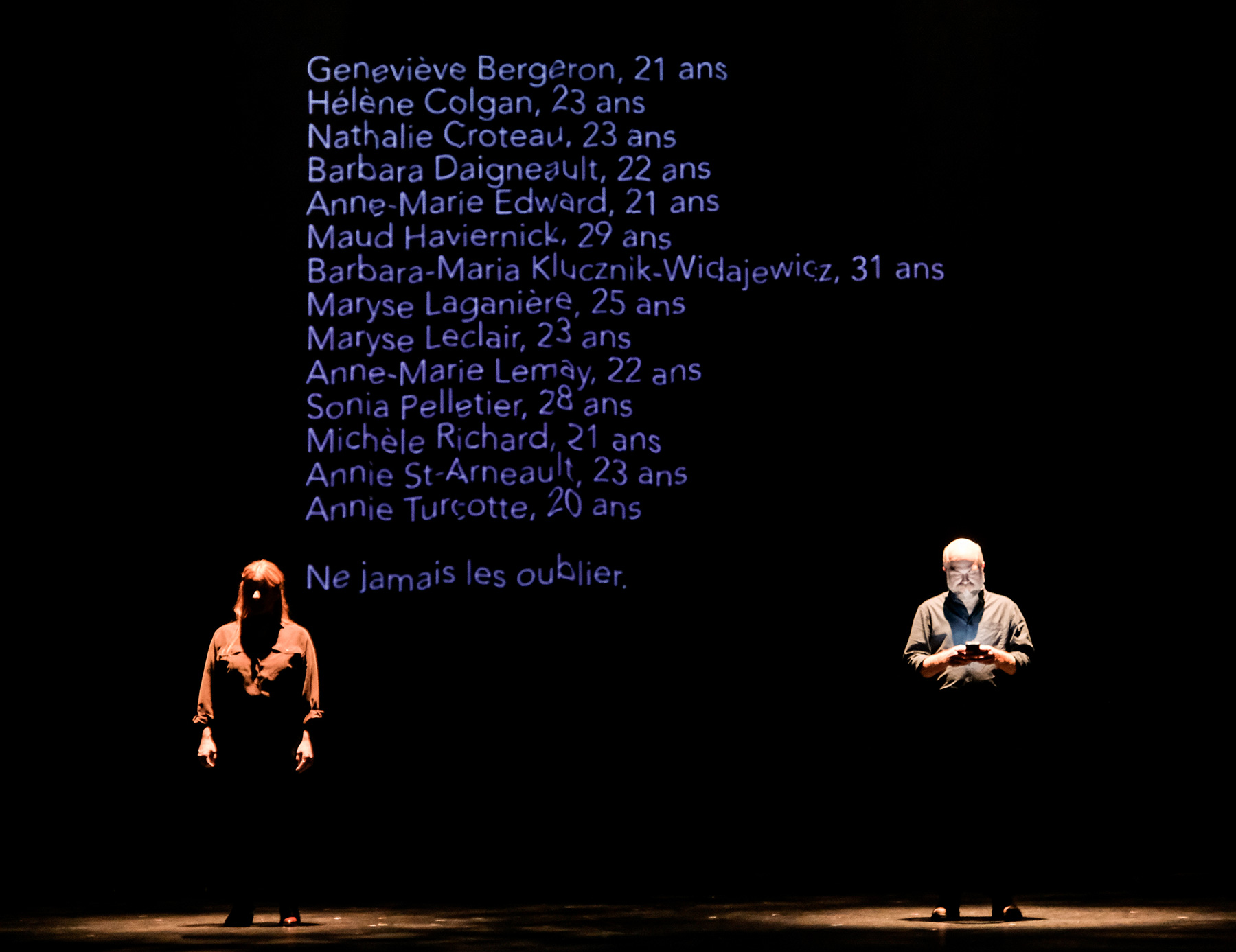 Photo of a man and woman standing under spotlights on a dark stage. Projected behind them are the names and ages of fourteen woman, the victims of the École Polytechnique massacre.
