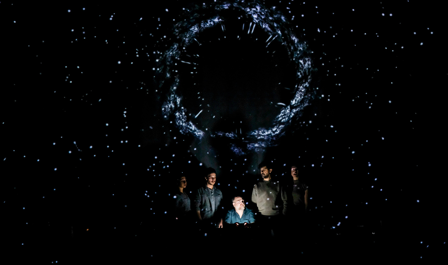 Photo of a group of people in shadow on a dark stage. Projected above them is a circle of light and scattered stars.