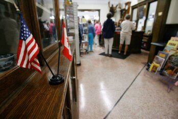 A black line across a library floor. In the foreground on a wooden shelf are small American and Canadian flags. Library patrons are seen in the background.
