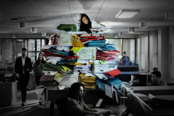 A photo of a stressed woman on top of a mountain of paperwork in the middle of her office.