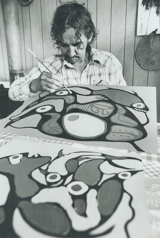 A black-and-white photo of Norval Morrisseau in a plaid shirt, looking down at a painting he is working on, a thin paintbrush in his hand. He is angling the painting up towards his face.
