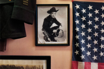 A photo hangs on the wall of a man with a cowboy hat and boots, holding a whip. On the wall also hangs an American flag, and what appears to be a military jacket.
