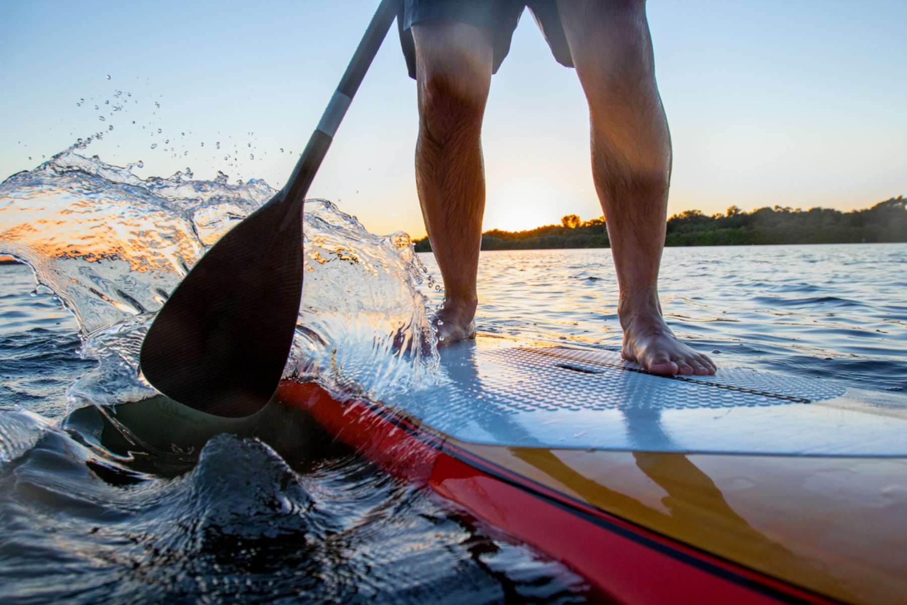 A close up of man on a paddleboard at sunset. He's pushing the paddle forward with water spraying forward to the camera
