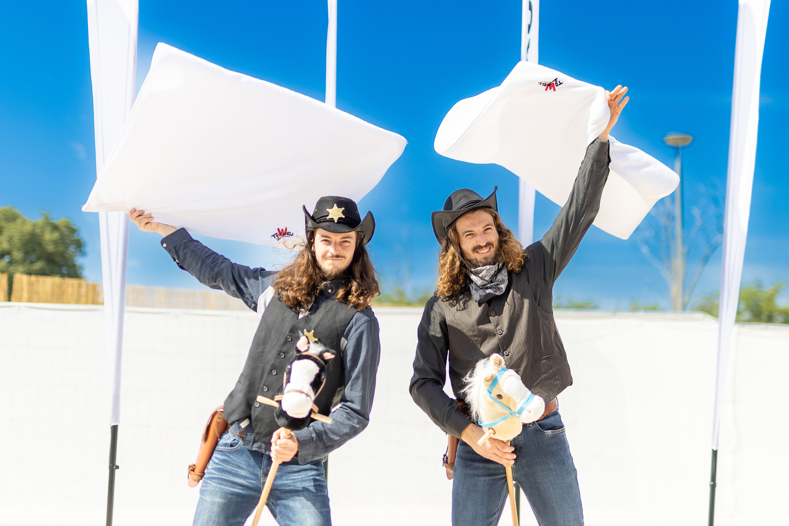 Photo of two men dressed as cowboys riding hobby horses while spinning towels in the air.