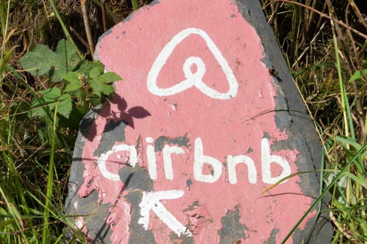 Photo of a rock painted pink with the Airbnb logo and company name painted on it in white sitting in the grass.