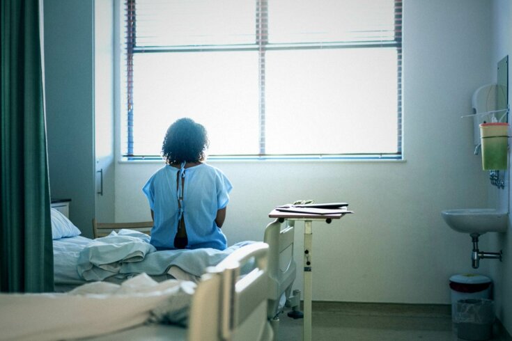 A photo of the back of a lonely woman in a hospital gown sitting on a bed while looking out of the window of her hospital room.