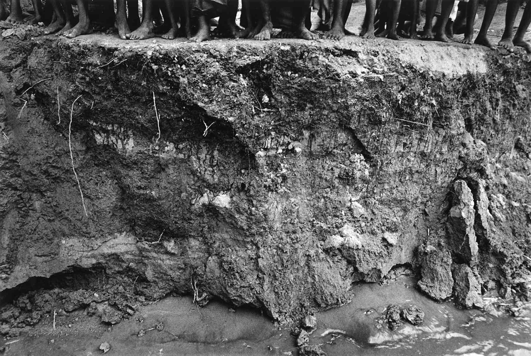 In a black-and-white photo, a row of people's feet can be seen standing on the bank of an eroding river bank.