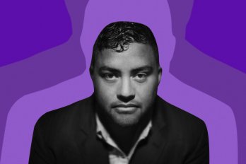 A photo of the poet, Shane Book, who stares at the camera with a serious expression. Behind him is a purple background in three shades, from light to dark.