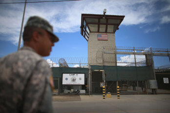 A photo of a soldier looking facing a tall fence and watch tower of a prison. The sky is a bright blue with white clouds