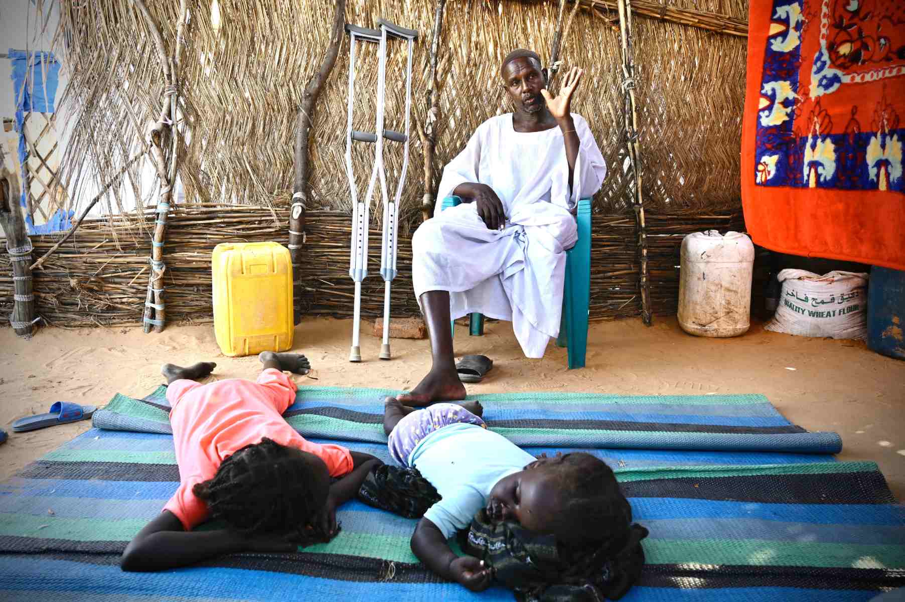 Photo of a man with one leg sitting in a chair with crutches nearby. Two young children sleep on a mat on the floor in front of him.
