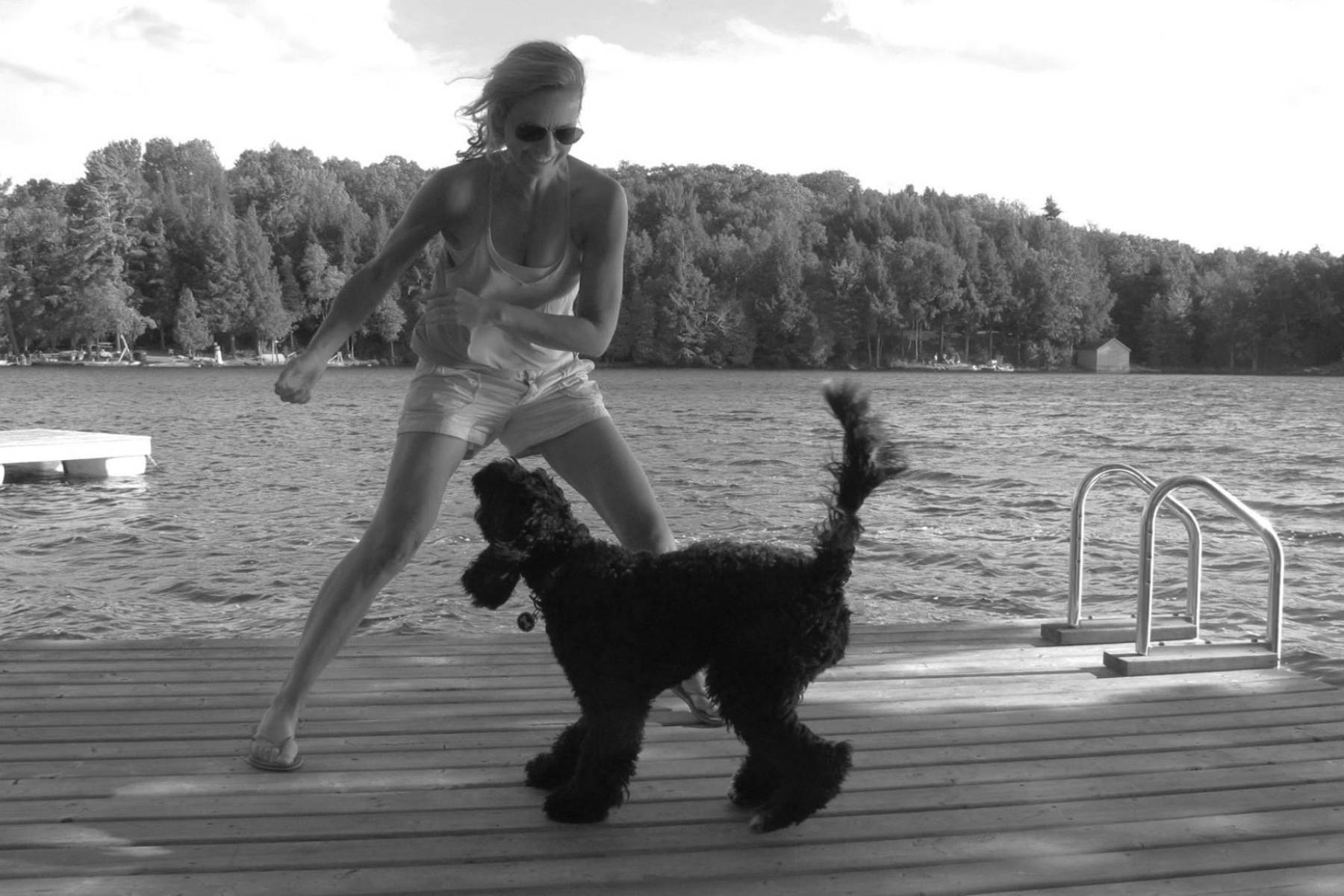 Black and white photo of a woman on a wooden dock in shorts, a tank top, and flip-flops playing with a black dog.
