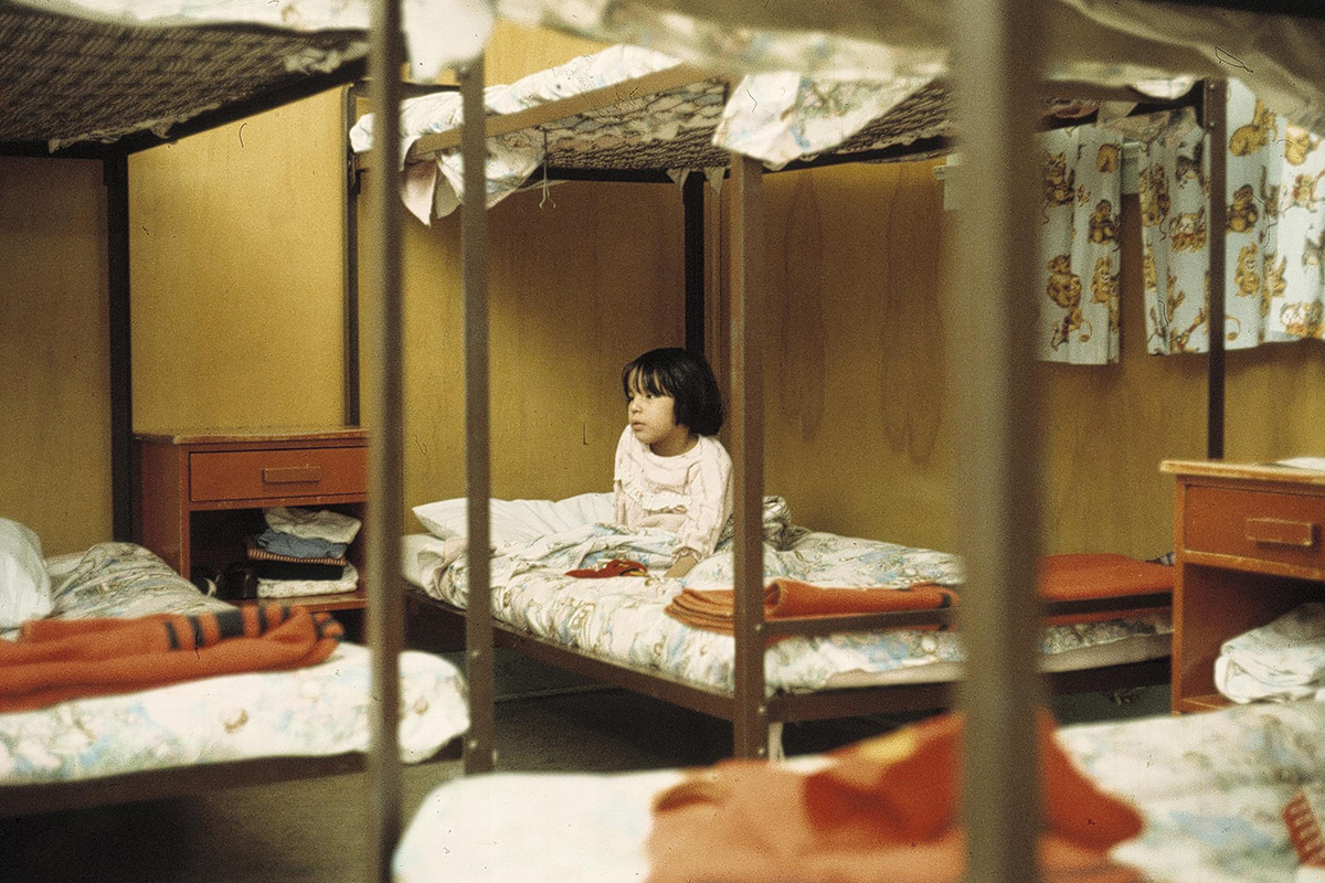 Photo of a young girl with short, dark hair. She is wearing pajamas and sitting up in the bottom bunk of a bunk bed, in a room with several other bunk beds.