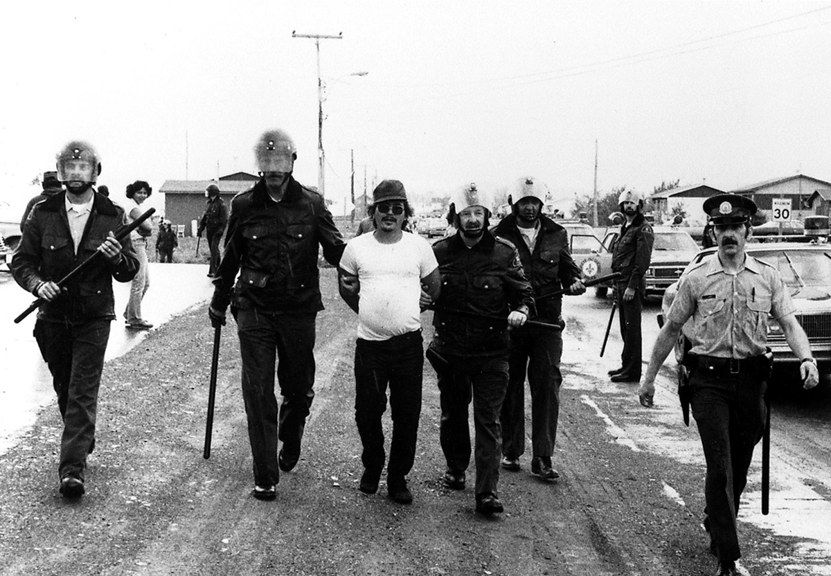 Black and white photo of a man wearing a white t-shirt, sunglasses, and baseball cap. His hands are behind his back and two police officers are holding his arms, leading him down a dirt road. Several other police officers are visible, some wearing helmets with visors and carrying batons.