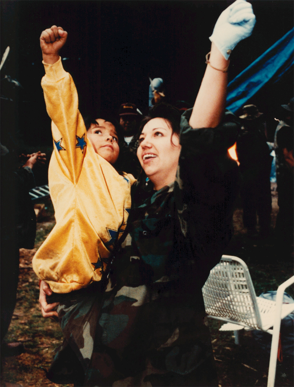 Photo of a woman holding a young child in her arms. They are both raising an arm, their hands in fists, and looking up. The woman is wearing camo print and the child is wearing a bright yellow sweater with blue stars.