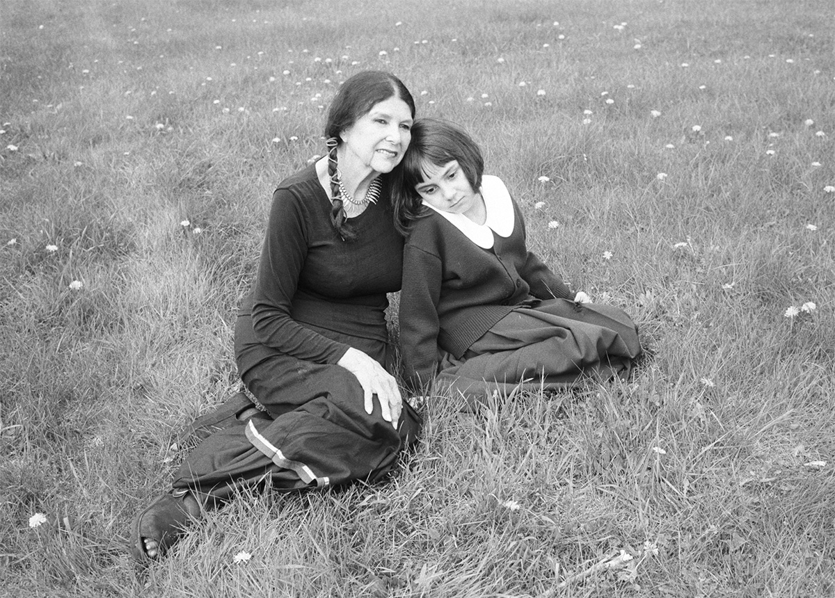 Black and white photo of a woman (Alanis Obomsawin) and a young girl sitting together in a grassy field. Alanis's hair is tied in braids and she is wearing a dark dress with long sleeves. The girl has short dark hair and she is wearing a skirt and sweater with a large white peter pan collar.
