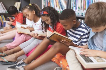 A row of children sit in front of a bookcase and read books studiously.
