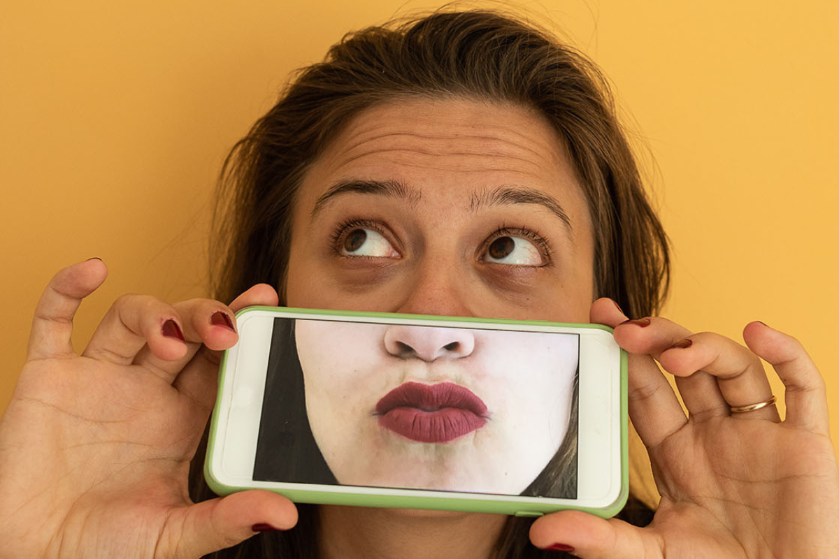 A woman wrinkles her brow and holds up a phone with a photo of her mouth over where her real mouth would be.