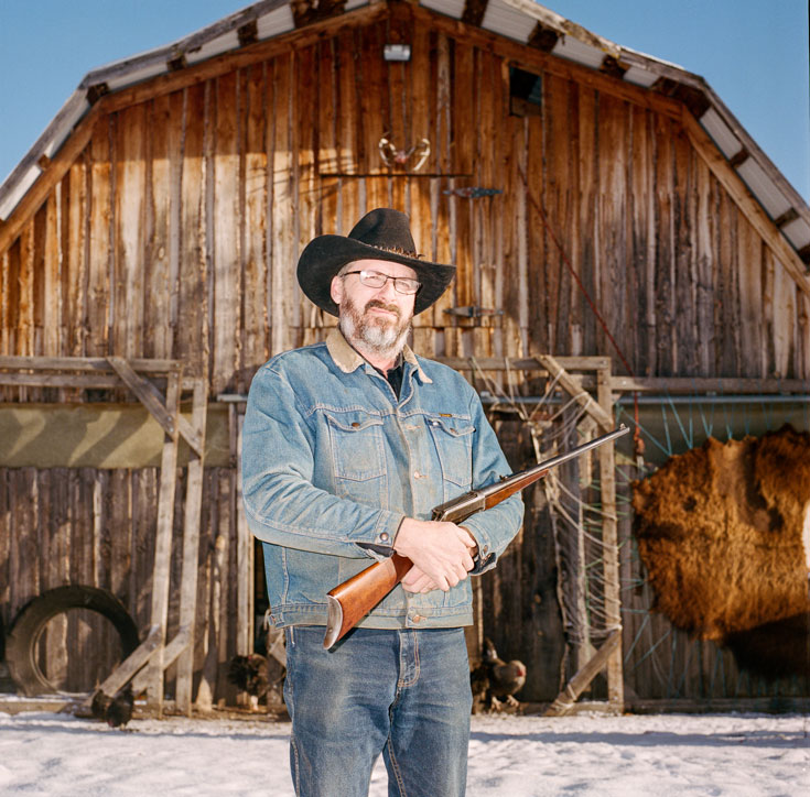 A bearded man stands in front of a barn and carries a rifle. He is wearing blue denim and a brown cowboy hat.
