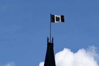 Against a blue sky, a black and white flag flies from the very top of the Parliament building. Instead of a Canadian flag, it's black and white with an oil drum replacing a maple leaf