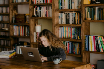 A young girl sits in front of a laptop at a wooden table. Behind her are shelves lined with books