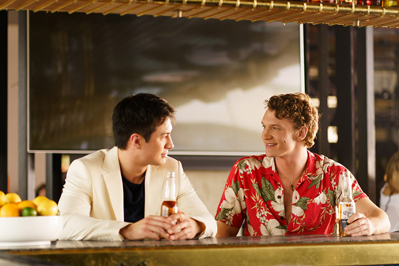 A man in a white blazer speaks to a man in a red Hawaiian shirt at a bar.