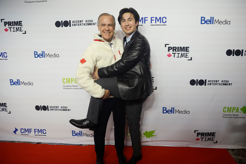 A photo of Hudson Williams with his leg up on Mark Carney, who is wearing a white fleece sweater