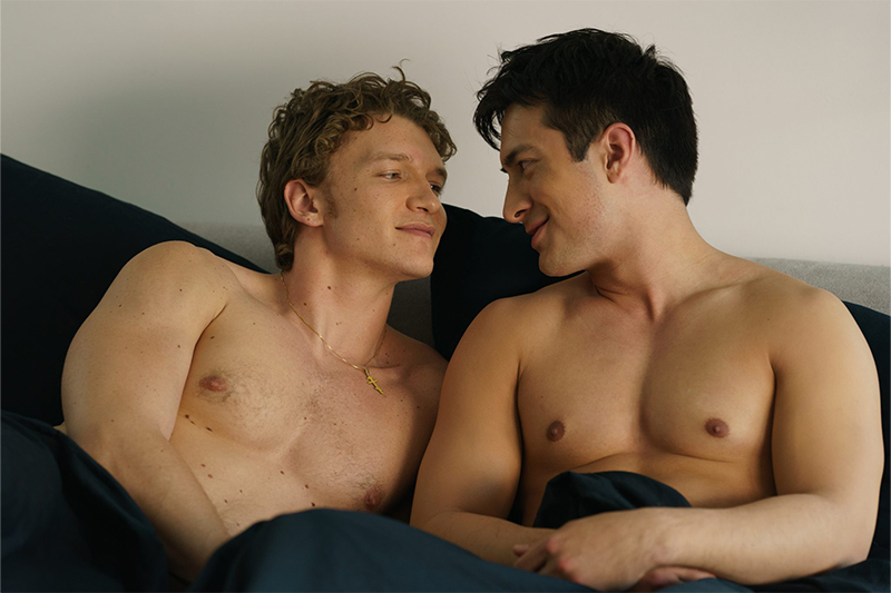 Two shirtless men in bed look at each other lovingly.