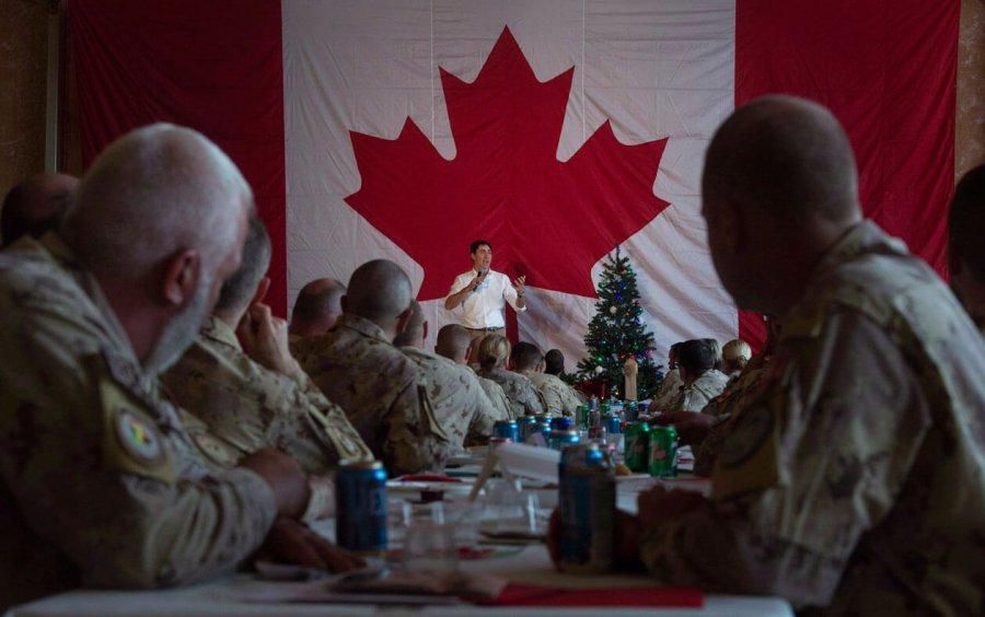 Justin Trudea stands in front of a Canadian flag and beside a Christmas tress while soldiers watch, seated.