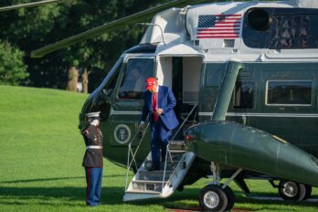 A photo of Trump, in suit and red MAGA cap, descending the Marine One helicopter as a man saltues
