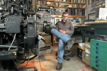 Photo of Gaspereau Press co-founder Andrew Steeves seated at his desk in his office, legs crossed, with a wall of books and binders behind him and a desk and printing equipment on either side of him.