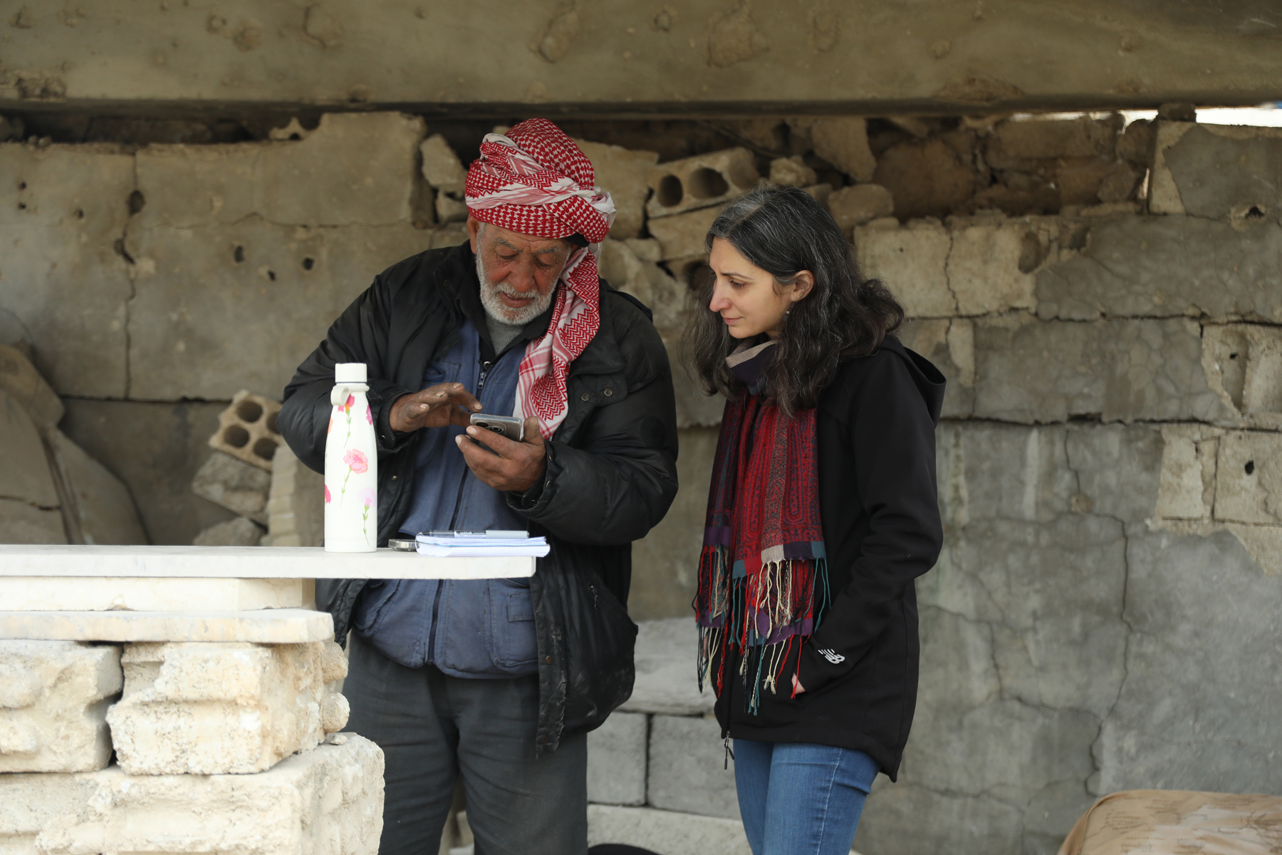 A photo of a man and a woman dressed in jackets, looking at a phone. Behind them and in front of them are cinderblocks
