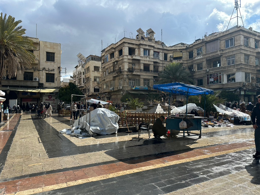 A photo of a square surrounded by mid-rise building. There is a blue umbrella in front of a table