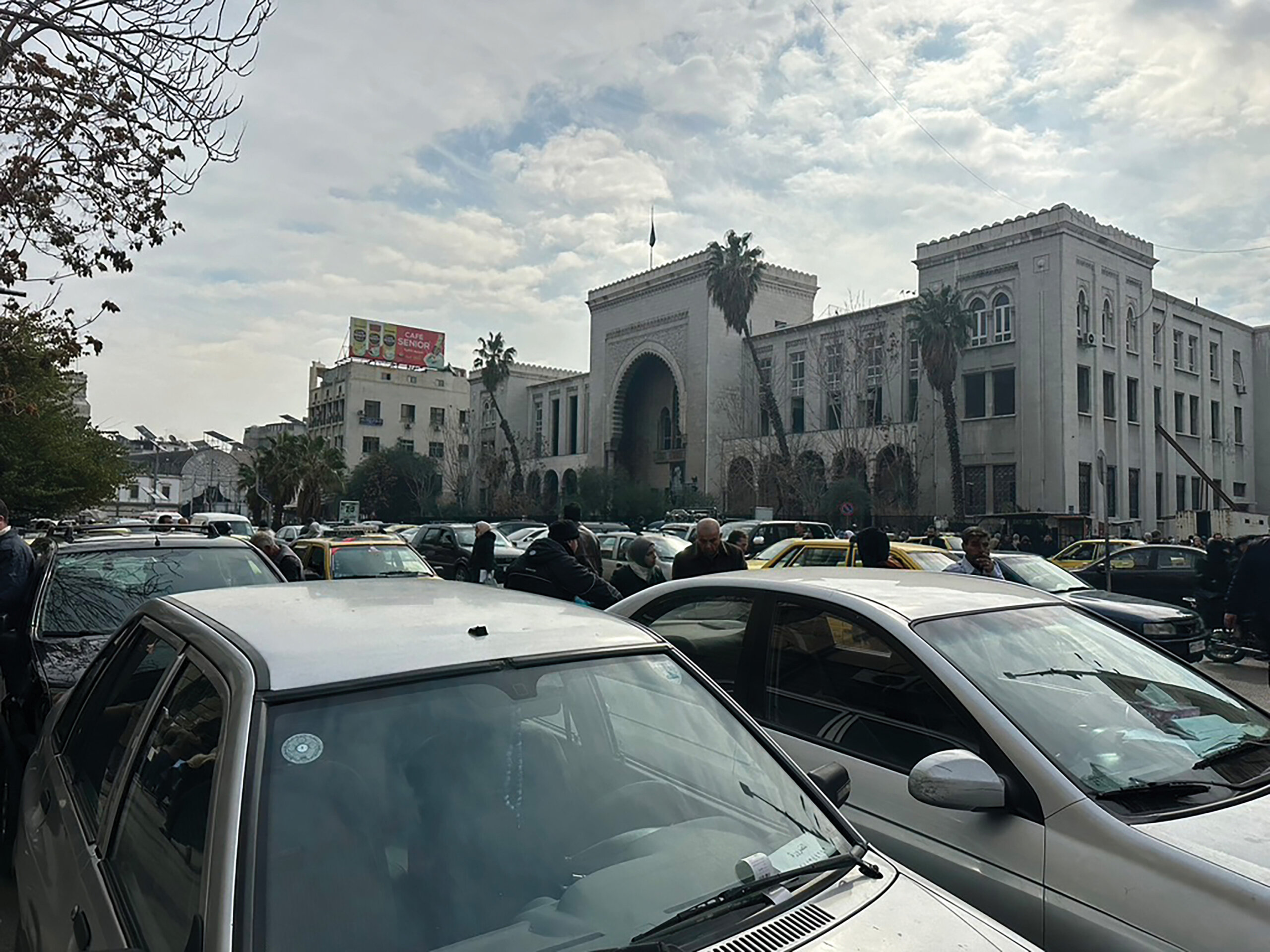 A photo of a large building with a grand, arched entrance, flanked by large palm trees. Cars speed past it on the busy street.