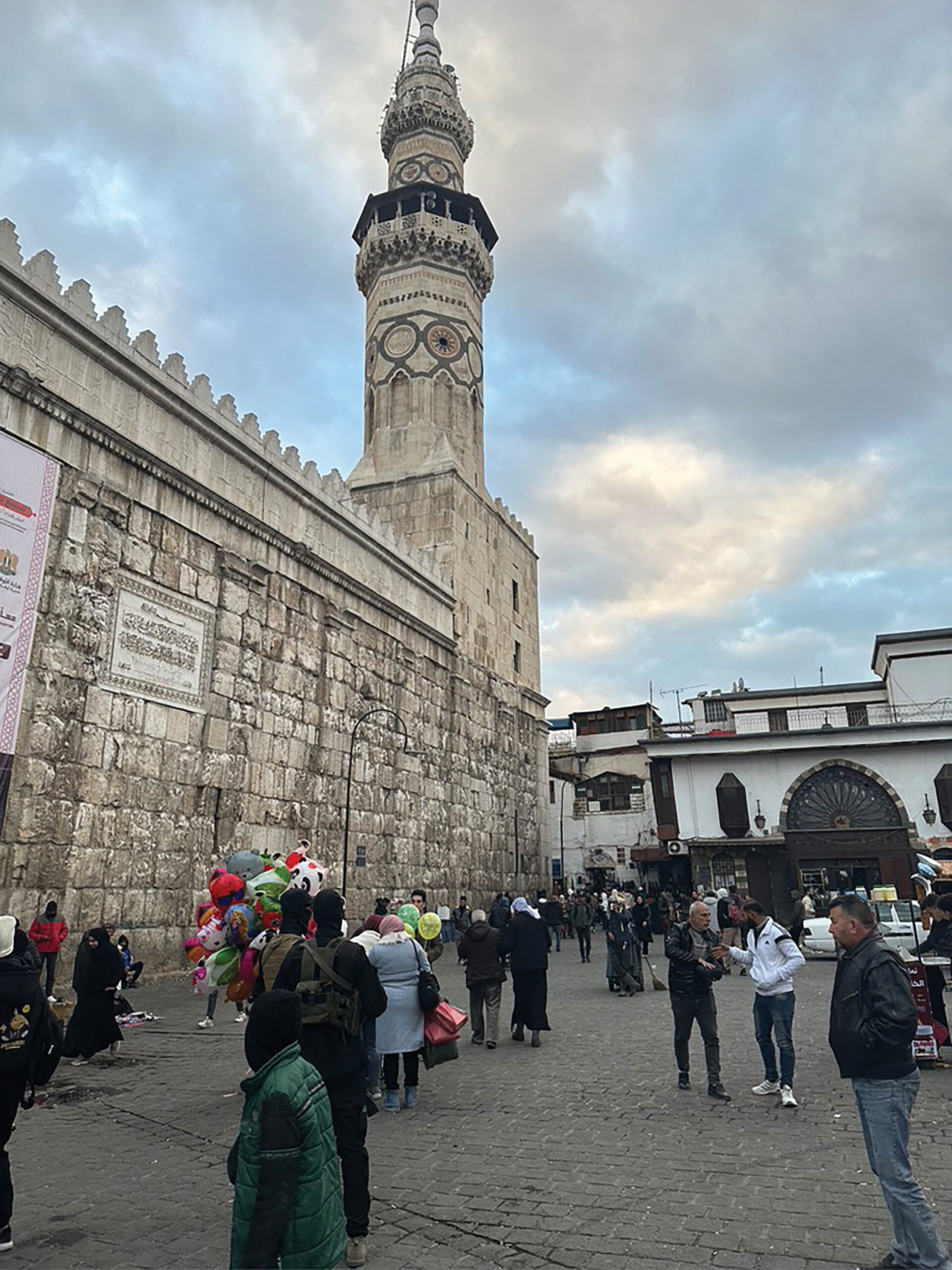 A mosque minaret. People mill about in the street below including a few who hold colourful balloons.