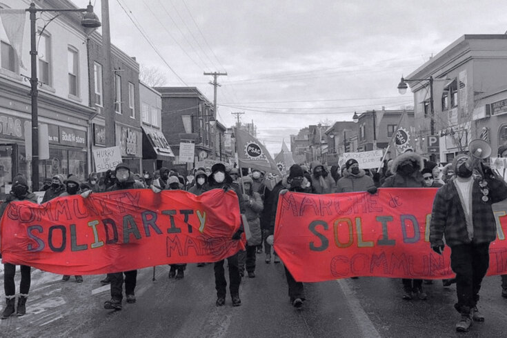 Photo of marching protesters. The people and scenery are in black and white, and the banners they are holding are in colour. The banners are red and read "Community Solidarity March."