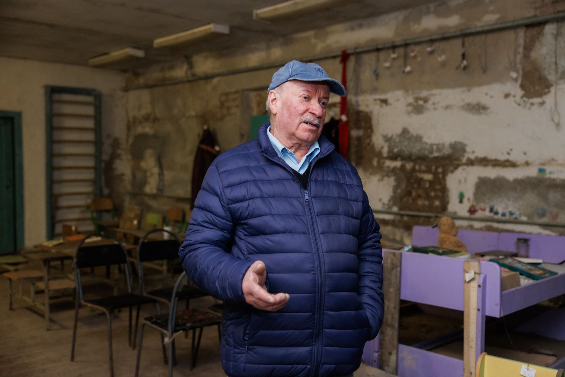 A photo of an older man, wearing a cap and blue coat, standing in a basement and gesturing with one hand