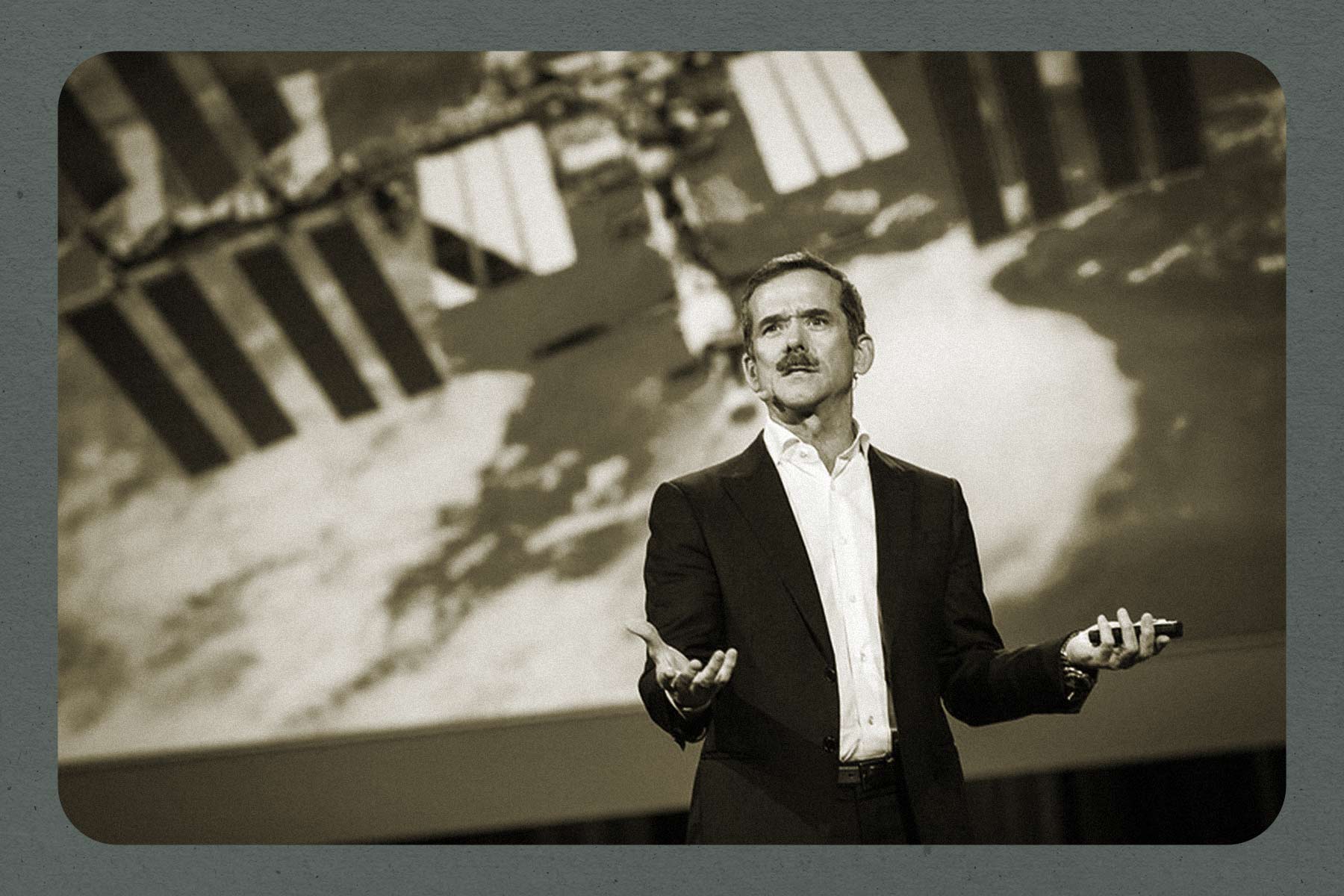 A black-and-white photo of Chris Hadfield speaking at a TED Conference in 2014. There is a blue border around the image.