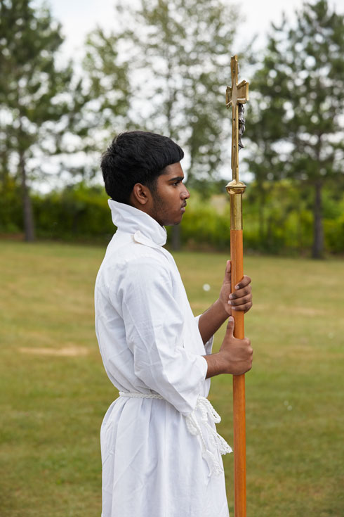 A young man in a white robe carries a large cross and faces the right of the frame.