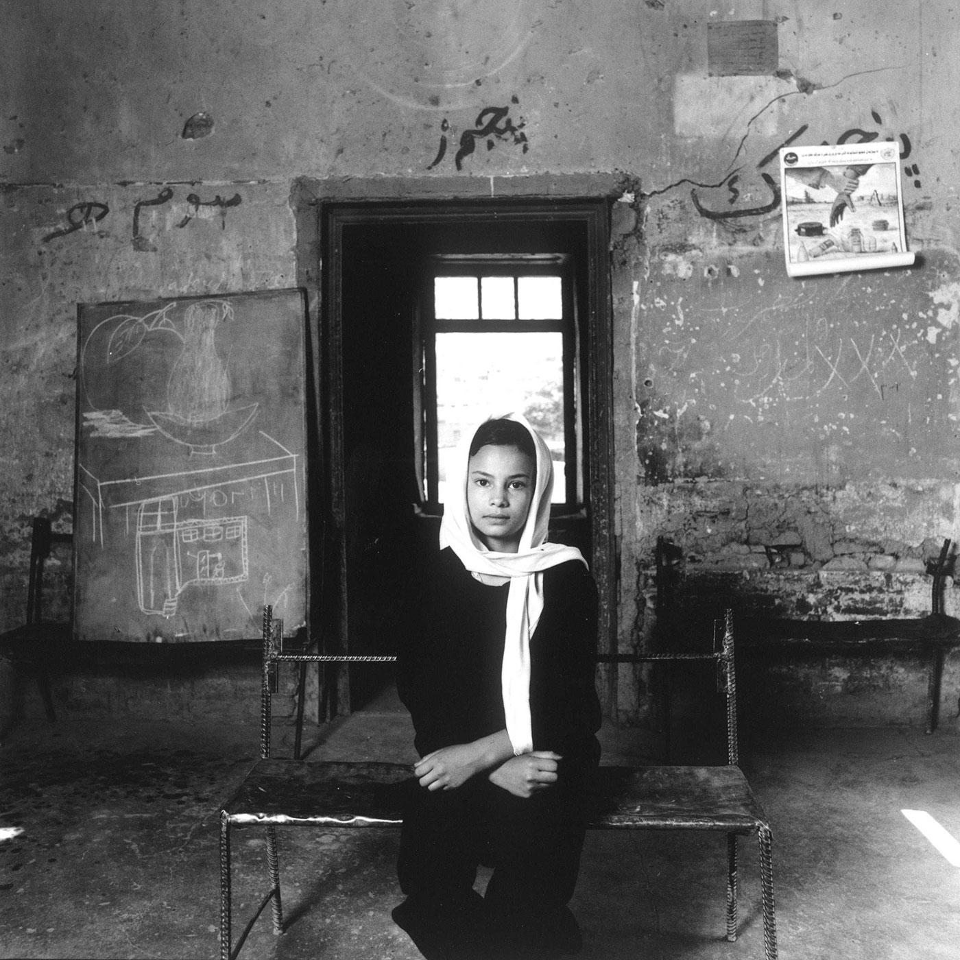 Black and white photograph of a young girl wearing a loose head scarf and dark clothing sitting on a metal bench in a classroom with stone walls.