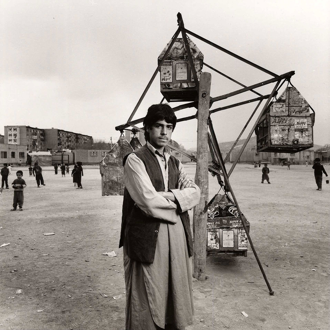 Black and white photograph of a young man, standing outside with his arms crossed, in front of a small Ferris wheel made of wood and scrap metal.