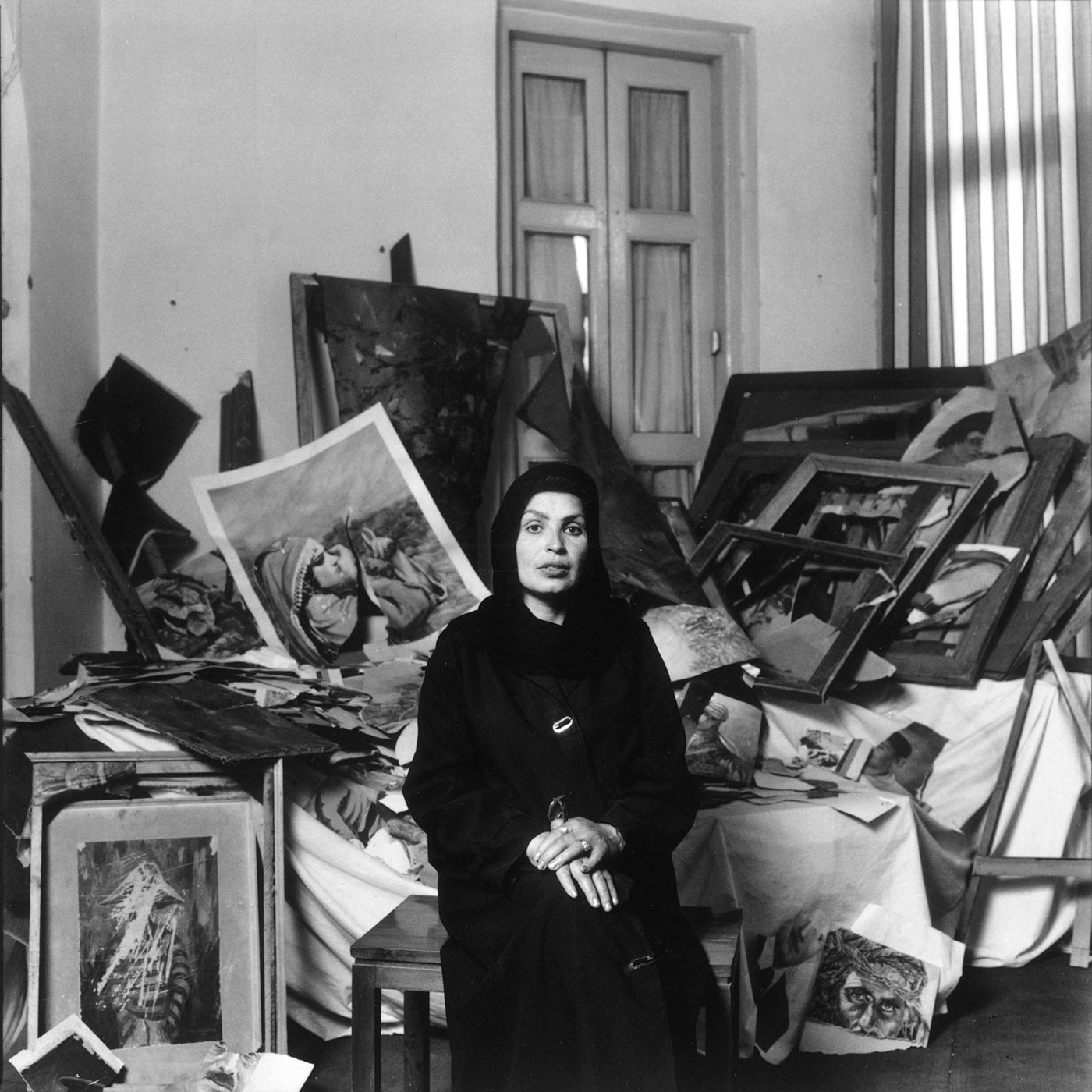 Black and white photograph of a seated woman wearing a head covering and dark clothing. Behind her is a tall, disorganized heap of damaged artwork and empty frames.