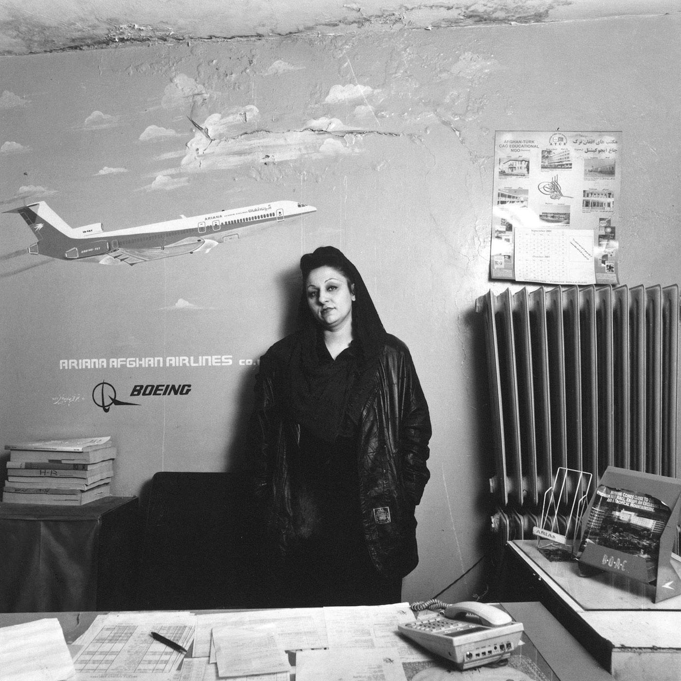 Black and white photograph of a woman wearing a head covering and leather jacket over dark clothing. She is behind a desk covered in paperwork and in front of a wall painted with artwork of an airplane in the sky.