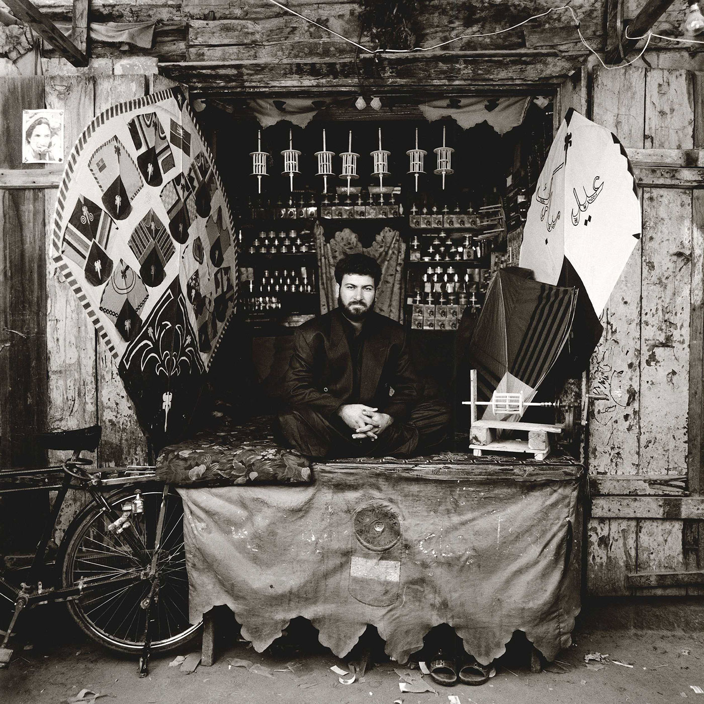 Black and white photograph of a man wearing a dark suit. He is sitting in a market stall decorated with kite-making tools and designs.