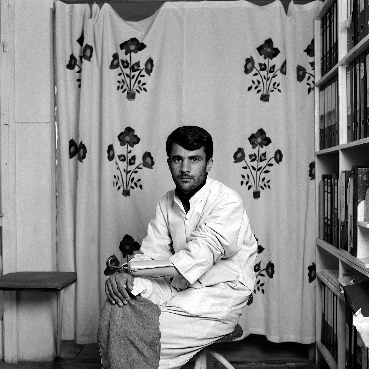 Black and white photograph of a seated man wearing light clothing, a sleeve rolled up to reveal a prosthetic arm with a hook on the end. He is in a room with a bookcase and a patterned curtain.
