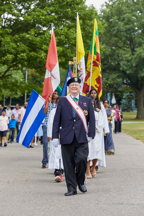 The procession carries flags and follows a smiling older man in a Knights of Columbus uniform.